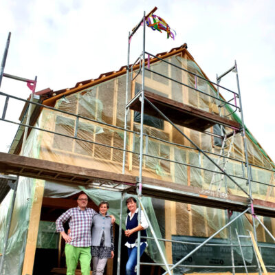 Picture of a LORENZ reference with the client couple and the planner in front of an albgeplanten straw construction on the scaffolded house with topping-out wreath