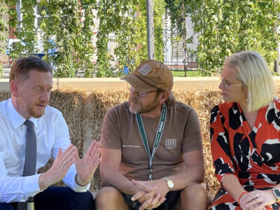 Picture of a conversation between Saxony's Minister President Michael Kretschmer with LORENZ founder Rainer K. Schmidt and MP Christiane Schenderlein on the occasion of the North Saxony Volunteer Festival at the Ur-Krostitzer brewery on a straw couch made from LORENZ wood and straw assembly systems made from wood and straw.